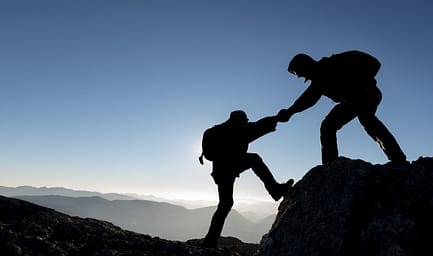 a silhouette of a man helping another man climb a rock